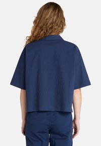 Camicetta corta a maniche corte di colore blu navy con una sottile texture stropicciata e colletto classico. Design pulito senza loghi o motivi visibili.