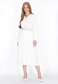 Weißes langärmeliges Kleid mit gerafftem Ausschnitt und silbernem Taillenband. Fließendes Design, knielang, glatte Stoffqualität, kombiniert mit weißen Absätzen.
