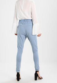 Pantalons bleu clair avec une taille élastique et une coupe fuselée, associés à un chemisier blanc avec des manches à volants en forme de cloche. Chaussures à talons noires.
