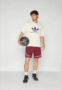 Jaunas vyras laiko juodą krepšinį, vilkėdamas off-white spalvos Adidas krepšinio marškinėlius, bordo spalvos šortus, baltas kojines ir juodus sportbačius.