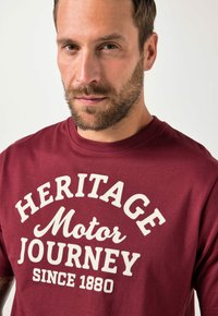 Maroonfarbenes Baumwoll-T-Shirt mit weißem Schriftzug: "HERITAGE Motor JOURNEY SINCE 1880." Rundhalsausschnitt und kurze Ärmel. Glatte Textur.