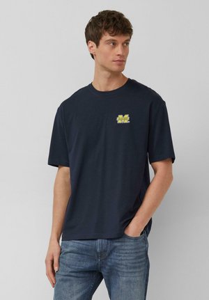 Młody mężczyzna w granatowej koszulce z małym żółtym logo University of Michigan na piersi i niebieskich dżinsach, stoi z ręką w kieszeni.