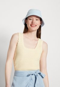 Top de ganga em croché amarelo claro com decote ondulado, combinado com um chapéu de balde azul claro. O top apresenta um padrão texturizado.