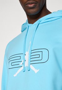 Svetlomodrá mikina s klokankou, kapucňou so šnúrkami a výrazným bielym logom znázorňujúcim postavu v skákajúcej póze s textom "23".