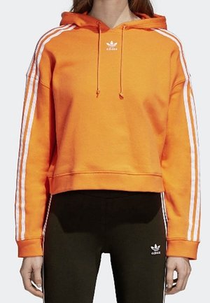 Osoba nosząca pomarańczową bluzę z kapturem Adidas z białymi paskami na rękawach oraz czarne legginsy Adidas z logo na górnej części uda.