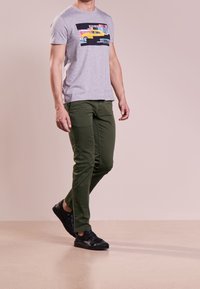 Grå kortärmad t-shirt med en färgglad grafik av en gul taxi; kombinerad med olivgröna byxor och svarta slip-on skor.