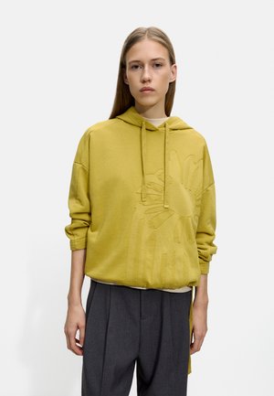 Żółta hoodie z kapturem i teksturowanym floralnym wzorem; ma luźny krój i ribbowe mankiety, w połączeniu z spodniami o wysokim stanie w kolorze szarym.