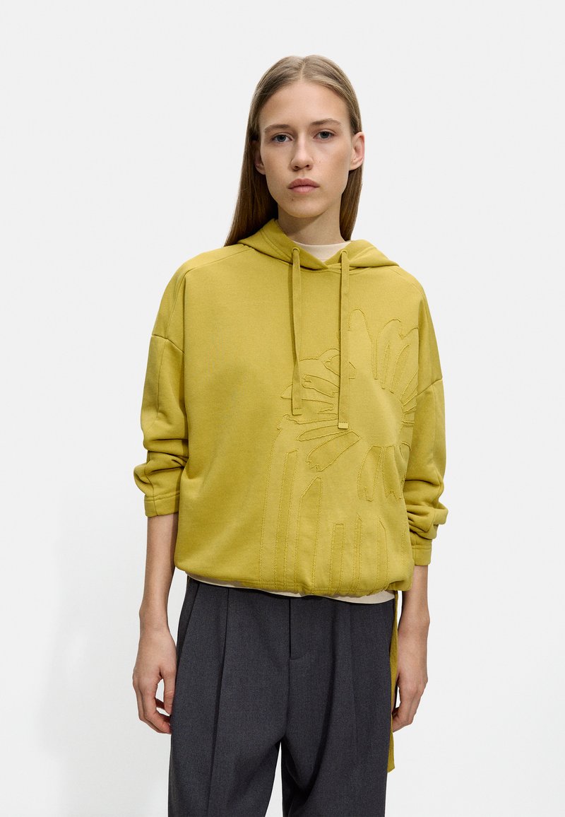 Żółta hoodie z kapturem i teksturowanym floralnym wzorem; ma luźny krój i ribbowe mankiety, w połączeniu z spodniami o wysokim stanie w kolorze szarym.