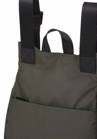 Delantal verde oscuro con anchas tiras negras ajustables en los hombros y un bolsillo frontal que contiene un objeto negro parcialmente visible.