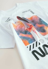 Tricou alb din bumbac, cu un grafic vibrant reprezentând o navetă spațială care urcă prin nori, cu text îndrăzneț și elemente de design în negru.