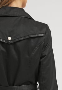 Czarna kurtka ze sztucznej skóry z kołnierzykiem, srebrnymi zatrzaskami oraz ściągniętą talią o gładkiej fakturze.