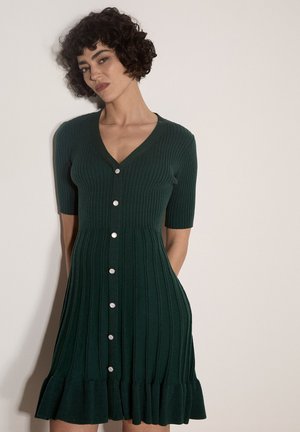 Donna con capelli corti, ricci e scuri, che indossa un vestito verde scuro a costine in maglia con maniche corte e bottoni frontali, in piedi davanti a uno sfondo chiaro e semplice.