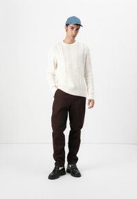 Crème gebreide trui, donkerbruine broek en zwarte loafers. Een blauwe pet maakt de look compleet. De minimalistische achtergrond benadrukt de outfit.