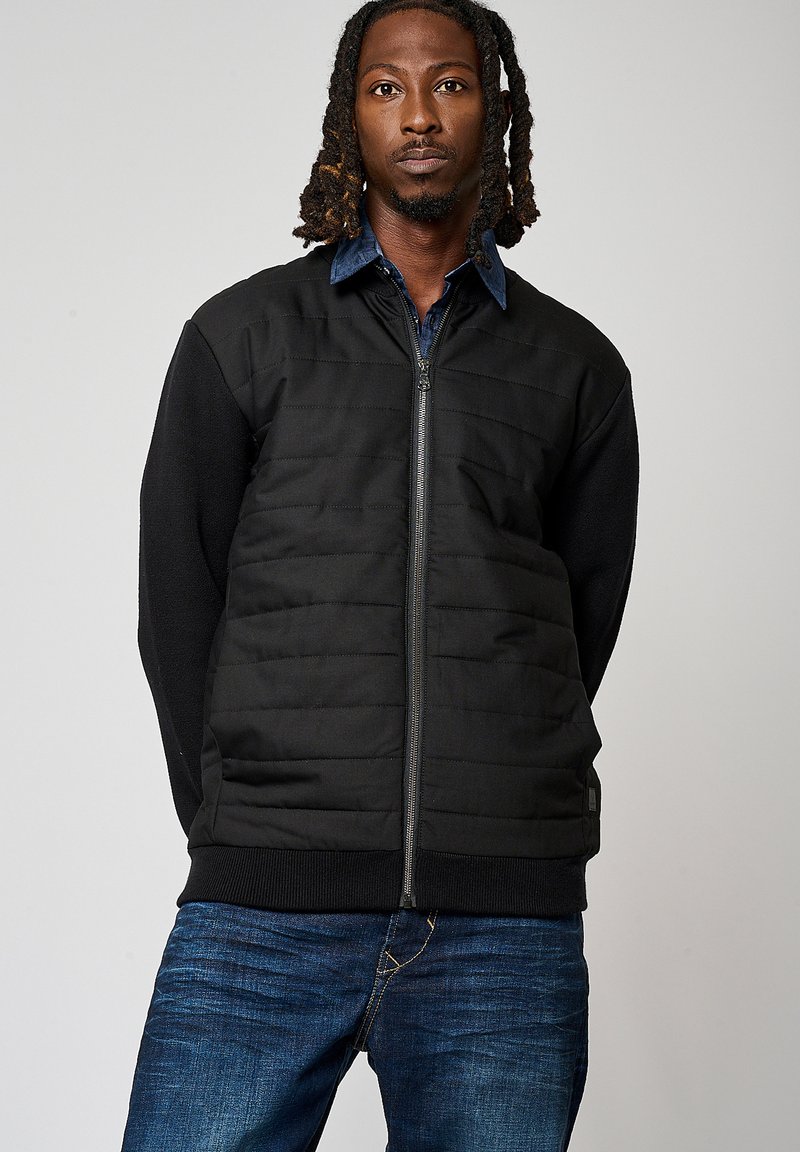 Veste isolante noire avec une fermeture éclair sur le devant, présentant un design matelassé sur le haut du corps et des poignets côtelés, associée à un jean en denim bleu.