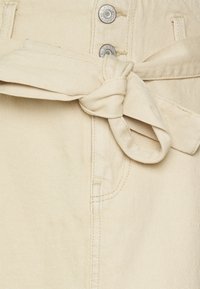 Tessuto di denim beige con dettaglio della cintura legata, chiusura a bottone argentato e cuciture visibili lungo le cuciture e i bordi delle tasche.