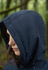 Marine waterdichte capuchon met zichtbare waterdruppels, voorzien van een trekkoord en een gladde textuur, geframed tegen een vervaagde groene achtergrond.