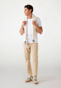 T-shirt blanc, pull en maille torsadée crème posé sur les épaules, pantalon beige et baskets marron clair avec des accents verts, debout devant un fond neutre.