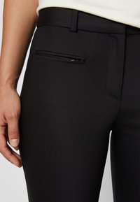 Zwarte broek gemaakt van een gladde, rekbare stof, met een ritszak aan de zijkant en een aansluitende pasvorm.