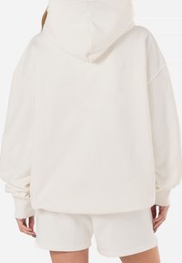 Sweat à capuche oversize couleur crème avec une poche avant, une capuche et des poignets côtelés, assorti à un short. Tissu doux et texturé.