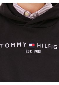 Col de chemise rayée boutonnée sous un sweat à capuche noir avec "Tommy Hilfiger" et logo de la marque brodés au-dessus de "EST. 1985".