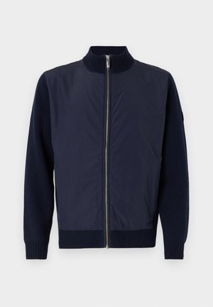 Giacca navy con colletto alto, dotata di una cerniera frontale, polsini in maglia e maniche in lana a contrasto per un tocco di texture in più.