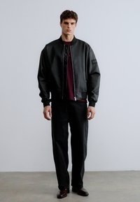 Juoda bombonešio stiliaus striukė su blizgiu paviršiumi, užsegama užtrauktuku, turinti ribuotas rankogalius, dėvima ant bordo spalvos marškinio. Derinama su laisvais tamsiais kelnėmis ir rudais batais.