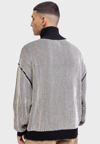 Pulover cu guler înalt, dungat în alb și negru, cu guler și manșete ribbed, croi oversized și țesătură texturată cu cusături vizibile.