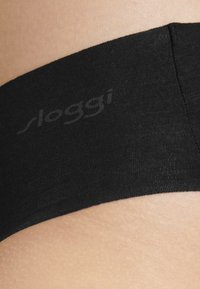 Schwarze Baumwollunterwäsche mit einem glatten, elastischen Bund und dem "sloggi"-Logo. Eine Nahaufnahme zeigt die weiche Textur und den Hautkontrast.