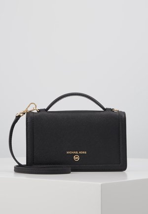 Sac à main rectangulaire noir Michael Kors avec poignée supérieure, bandoulière amovible et logo doré sur le devant, posé sur une surface blanche.