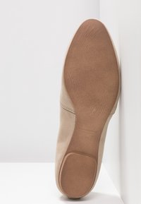 Semelle d'une chaussure plate en daim beige avec une semelle lisse couleur tabac et un talon texturé, positionnée verticalement contre un mur d'angle blanc.