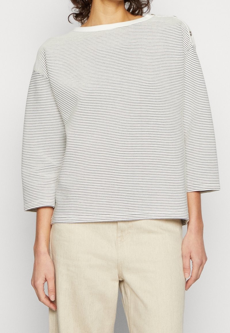Personne portant un haut blanc à manches longues avec de fines rayures horizontales noires et un pantalon beige clair, debout les bras détendus le long du corps.