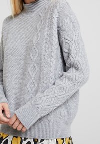 Světlý šedý svetr z pleteniny s vysokým límcem, vroubkovanými manžetami a texturovanými vzory na rukávech. Měkká tkanina z vlněného směsového materiálu.