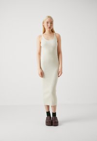 Abito midi senza maniche a costine di colore bianco sporco. Abbinato a zoccoli marrone scuro e calze verdi. Il modello è in piedi contro uno sfondo neutro.