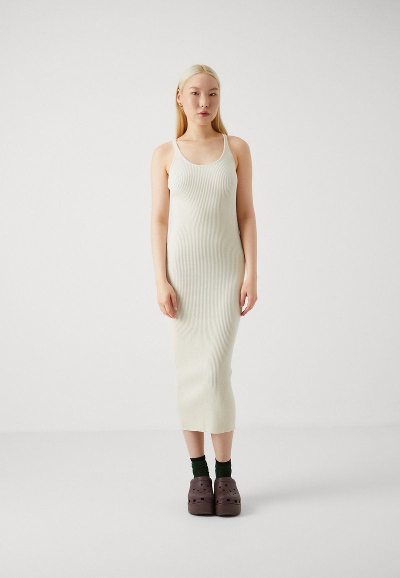 Abito midi senza maniche a costine di colore bianco sporco. Abbinato a zoccoli marrone scuro e calze verdi. Il modello è in piedi contro uno sfondo neutro.