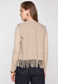Gilet beige lavorato a maglia con un motivo texturizzato e orlo a frange. Maniche lunghe e vestibilità comoda. Abbinato a jeans scuri.