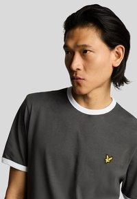 T-shirt de manga curta cinza escuro com colarinho e punhos brancos, apresentando um pequeno logótipo bordado amarelo no lado esquerdo do peito. Textura de tecido suave.