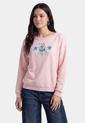 Donna che indossa una felpa rosa chiaro con un cane bianco e due stelle blu ricamate sul davanti, abbinata a jeans blu.