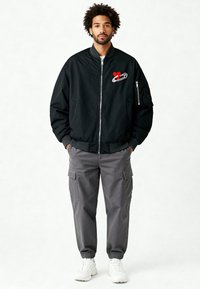 Veste bomber noire avec une broderie rouge en forme de cœur et une épingle de sûreté. Pantalon cargo gris et baskets blanches complètent le look.