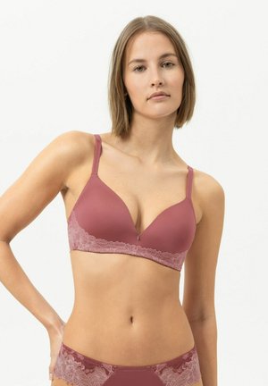 Frau mit kurzen Haaren, die passenden mauvefarbenen BH und Spitzenhöschen trägt, steht vor hellem einfarbigem Hintergrund.