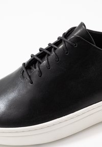 Zapatilla negra de cuero con una textura suave, forma redondeada y suela de goma blanca. Presenta cordones planos negros y un diseño minimalista.