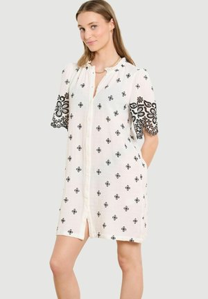 Mladá žena v bielych šatách na gombíky s čiernou kvetovanou výšivkou na rukávoch a tele stojaca pred svetlým jednofarebným pozadím.