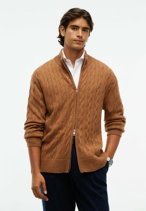 Mann trägt einen braunen, kabelgestrickten Zip-up-Pullover über einem weißen Hemd, mit dunkler Hose und Armbanduhr, steht mit einer Hand in der Tasche.