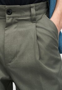 Κοντινή λήψη λαδί παντελονιού με μαύρο κουμπί και το χέρι μερικώς στην δεξιά τσέπη, που δείχνει την υφή του υφάσματος και τις λεπτομέρειες της ραφής.