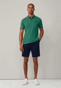 Grön pikétröja med marinblå detaljering vid kragen, kombinerad med marinblå shorts. Modellen bär vita sneakers. Enkel, vardaglig design. Inga mönster eller accenter.