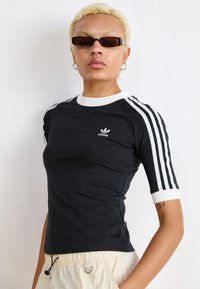 Svart tettsittende t-skjorte med hvit krage og tre hvite striper på ermene. Har Adidas-logo på brystet og en jevn tekstur.