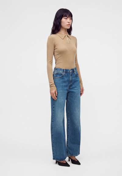 Femme debout portant un polo manches longues camel, un jean bleu à jambes larges et des talons noirs à bout pointu sur un fond blanc uni.