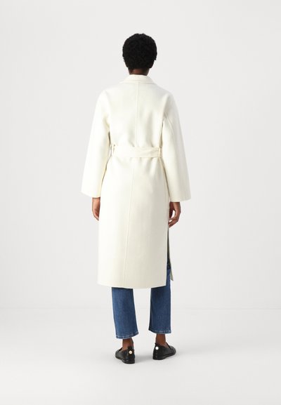 Weißer langer Mantel mit Gürtel, der eine glatte Textur, weite Ärmel und eine gerade Silhouette aufweist, kombiniert mit blauen Jeans mit weitem Bein und schwarzen Schuhen.