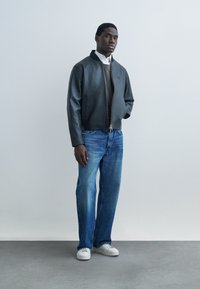 Homme debout portant une veste en cuir noire, une chemise blanche, un pull gris, un jean bleu et des baskets blanches, sur un fond clair uni.