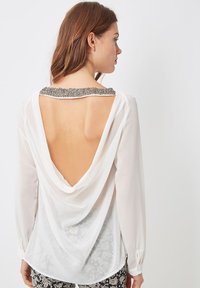 Mujer con cabello castaño, vistiendo una blusa blanca de manga larga con un escote de cuentas y una espalda baja, combinada con pantalones de estampado en blanco y negro.