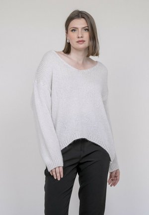 Jeune femme aux cheveux mi-longs portant des boucles d'oreilles en perles, un pull blanc en tricot ample et un pantalon noir, devant un fond clair uni.